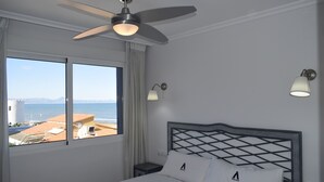 A hotel room with a bed, a ceiling fan, a nightstand, a lamp, and a window with a view of the sea and buildings. - Anacasa Medina Molins les Marines AP2201 (Denia)