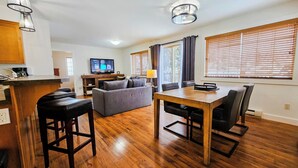 Una sala de estar moderna con un sofá gris, una mesa de comedor de madera y sillas negras. Un televisor de pantalla plana está montado en la pared, y hay una zona de cocina con armarios de madera y un taburete de bar.