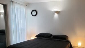 A bedroom with a large bed, a bedside table with a lamp, a wall clock, and a window with curtains. - Crescent Bar Condos Unit 143 (Quincy)