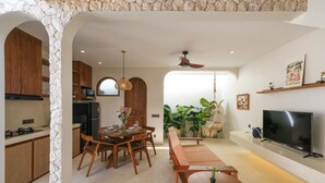 A modern kitchen with wooden cabinets, a dining area with a wooden table and chairs, a built-in TV, and a large window with plants. - Villa Koka Uluwatu (Pecatu)