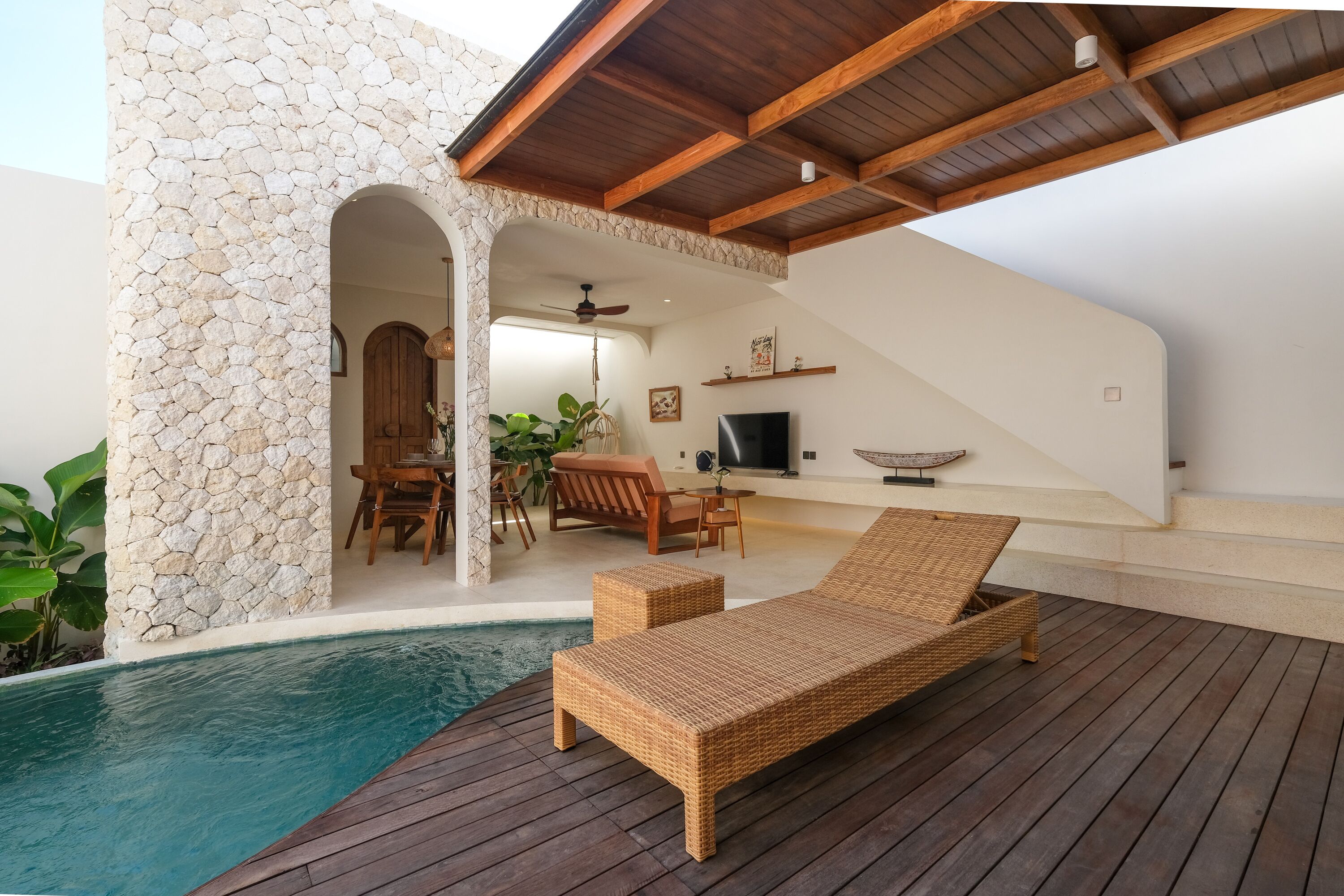 Un área junto a la piscina con una silla de mimbre, una pared de piedra y una terraza de madera.