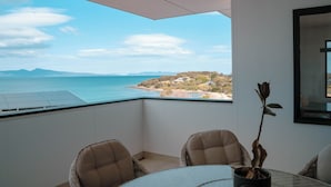 A balcony with a view of the sea and mountains, a table with chairs, and a potted plant.