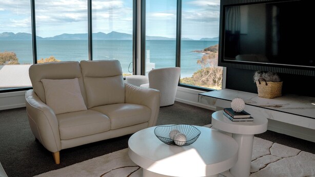 A modern living room with a white sofa, two armchairs, a round coffee table, and a large TV screen. The room features large windows with a view of the sea and mountains.