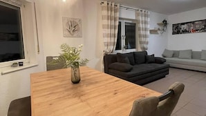 A dining area with a wooden table, chairs, and a vase of flowers. There is a black sofa and a white couch in the background.