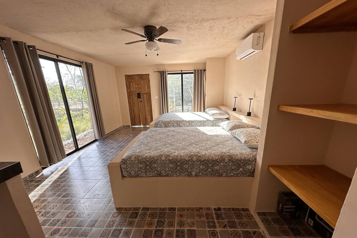 A bedroom with two beds, a ceiling fan, an air conditioning unit, and a view of greenery outside.