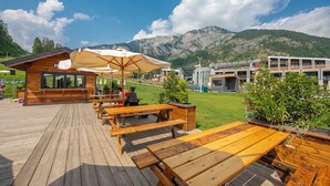 A wooden deck with picnic tables and umbrellas, a small building with a sign, and a mountainous backdrop. - The Secret Garden in the City center, Bardonecchia, Italy (Bardonecchia)