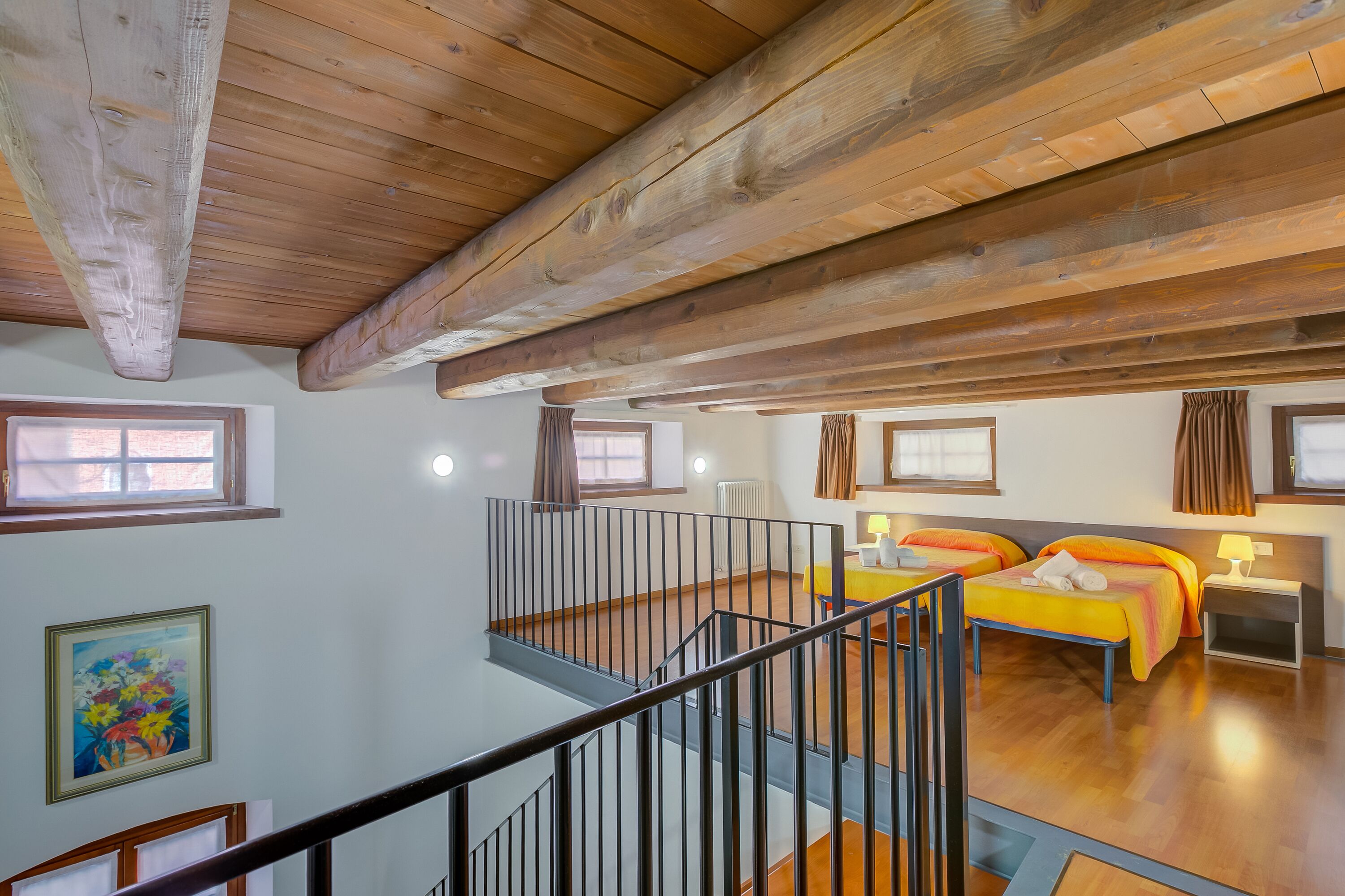 A spacious room with wooden beams on the ceiling, two beds with orange bedding, a small table, and a window with curtains.