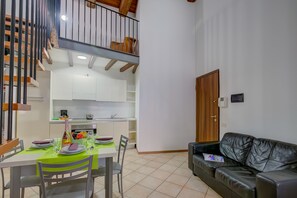 A dining area with a table set for two, a black leather sofa, a staircase, and a kitchen in the background.