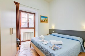 A hotel room with a bed, a window with curtains, a radiator, a painting on the wall, and a book on the bed.