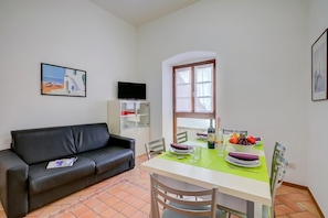 Un coin repas avec une table dressée pour deux personnes, un canapé en cuir noir, un téléviseur et une fenêtre avec des stores.