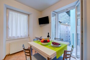 A dining area with a table, chairs, a bowl of fruit, a bottle, and a TV mounted on the wall.