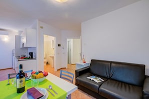 A modern living room with a black leather sofa, a dining table with a green tablecloth, and a kitchen area with white cabinets.
