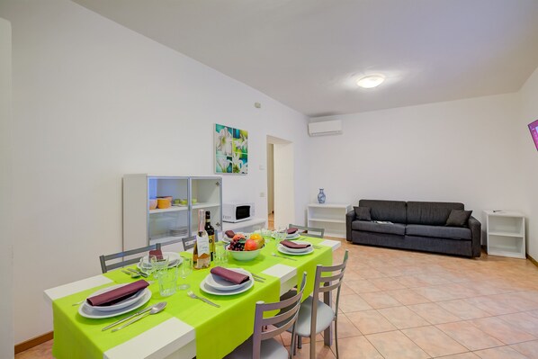 Zona pranzo con tavolo apparecchiato per quattro persone, un divano grigio e una parete bianca con un quadro.