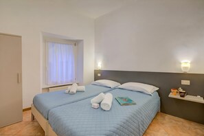 Un lit bien fait, avec une literie bleue, une fenêtre ornée de rideaux blancs, une table de chevet sur laquelle se trouvent un livre et une tasse, et un luminaire fixé au mur.