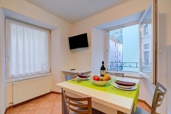 Un coin repas avec une table dressée pour deux personnes, un saladier de fruits, une bouteille de vin et un téléviseur fixé au mur.