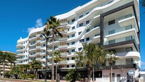 Un hôtel à plusieurs étages doté de balcons, de palmiers et surplombant un ciel d’un bleu éclatant.