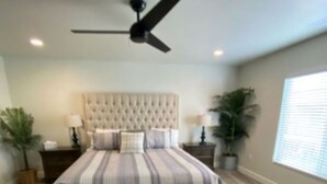 A bedroom with a tufted headboard, a bed with striped bedding, a ceiling fan, and two potted plants.