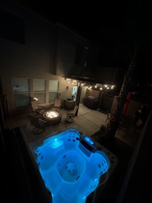 Un jacuzzi iluminado con luces azules, rodeado de mobiliario exterior y una chimenea.