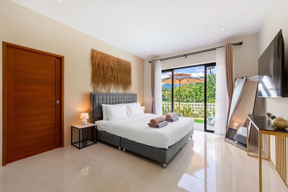 A modern bedroom with a large bed, a wooden door, a wall-mounted TV, and a balcony with a view.