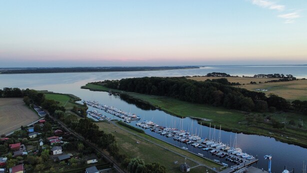 Une vue aérienne d’un port de plaisance où des bateaux sont amarrés le long d’un étroit canal, le tout étant entouré de verdure et de zones résidentielles.