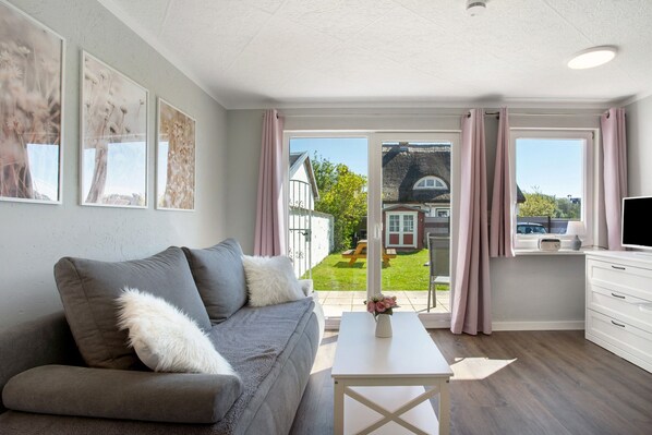 A modern living room with a grey sofa, white coffee table, and a view of a garden and house through large windows.