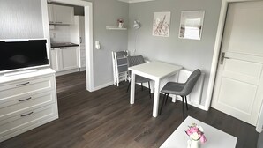 Una habitación moderna y bien iluminada con un televisor de pantalla plana, una mesa de comedor con sillas, una mesa de centro blanca con un florero y una zona de cocina con armarios blancos y una encimera blanca.