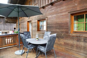 Une terrasse en bois avec une table ronde et des chaises, un parasol et une fenêtre donnant sur un paysage verdoyant.