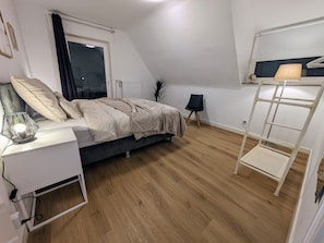 Ein Schlafzimmer mit Bett, Nachttisch, Lampe, Fenster, Heizkörper, Sessel und einem weißen Regal.