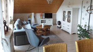 Ein Wohnzimmer mit Sofa, Sesseln, Fernseher, Schrank und Deckenleuchte.