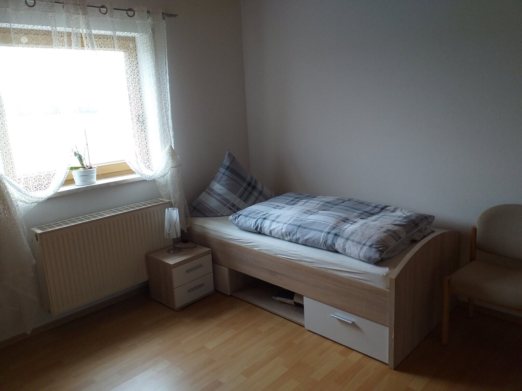 Une petite chambre avec un lit simple, une fenêtre, un radiateur, une table de chevet et une chaise.