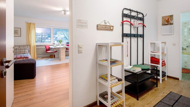 Une pièce comprenant un lit, une chaise rouge, une étagère blanche avec des livres, une étagère noire avec un coussin vert et une étagère noire avec un sac rouge.