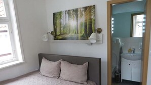 Une chambre à coucher avec un lit, un tableau représentant une forêt, une table de chevet et une salle de bain avec un lavabo et un miroir.