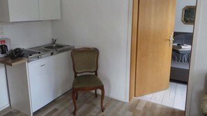 Une petite cuisine avec des meubles blancs, un évier et une chaise. Une porte en bois donne sur une chambre à coucher où se trouvent un lit et un tableau accroché au mur.