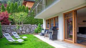 A modern house with a patio, two lounge chairs, a grill, and a stone wall.