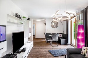 Uma sala de estar moderna com uma TV de tela plana, um espaço de refeições com mesa e cadeiras, e uma grande janela com cortinas.