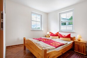 Une chambre à coucher avec un lit en bois, deux tables de chevet, une commode, deux fenêtres et une vue sur l’extérieur.