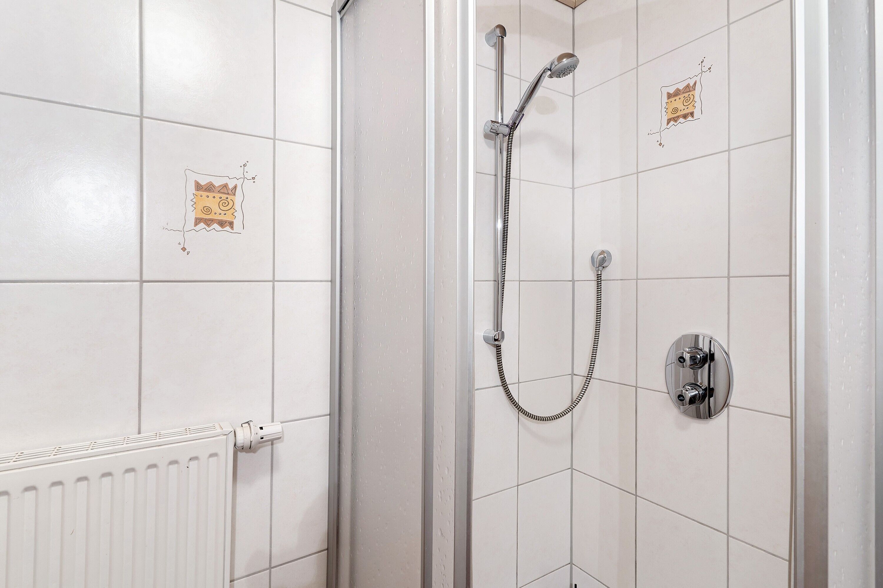 Une douche équipée d’un pommeau de douche à main et d’un robinet mural, avec des murs recouverts de carreaux blancs et une prise électrique visible.