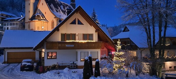 A two-story chalet with a balcony, snow-covered surroundings, and a mountain backdrop.