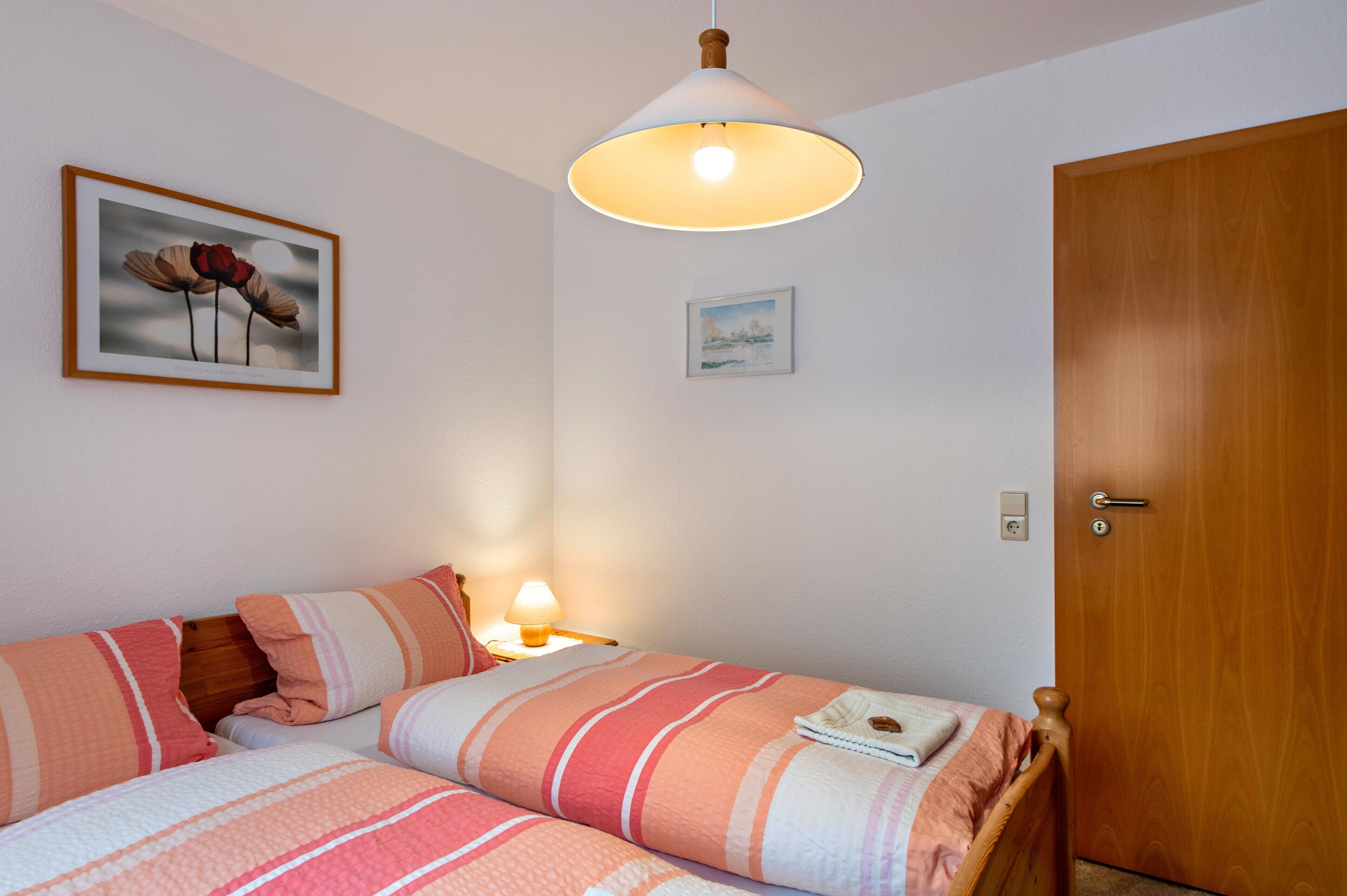 Un dormitorio con cama, lámpara de noche, puerta de madera y dos cuadros en la pared.