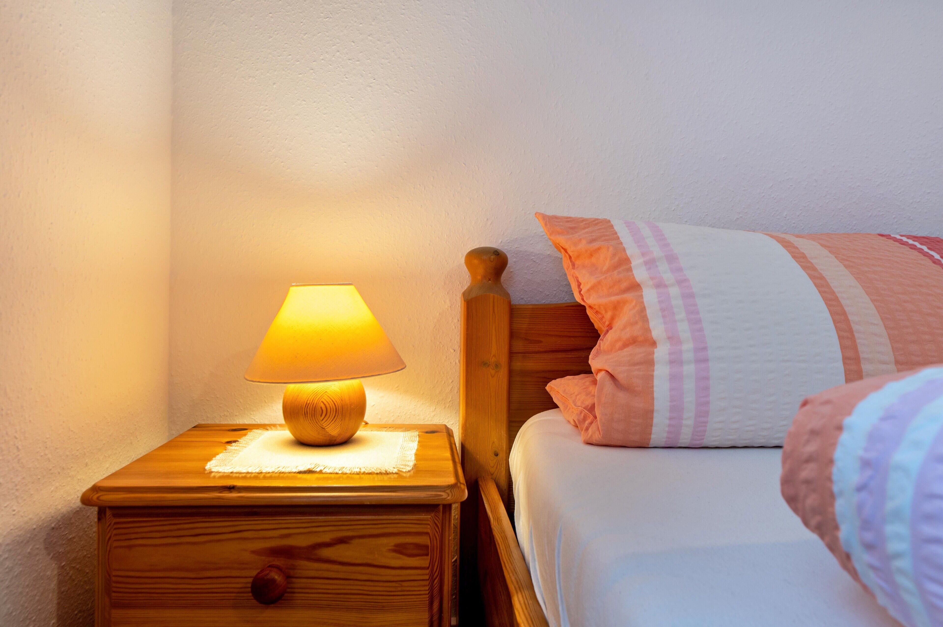 Een houten nachtkastje met een lamp, een bed met gestreept beddengoed en een witte muur.