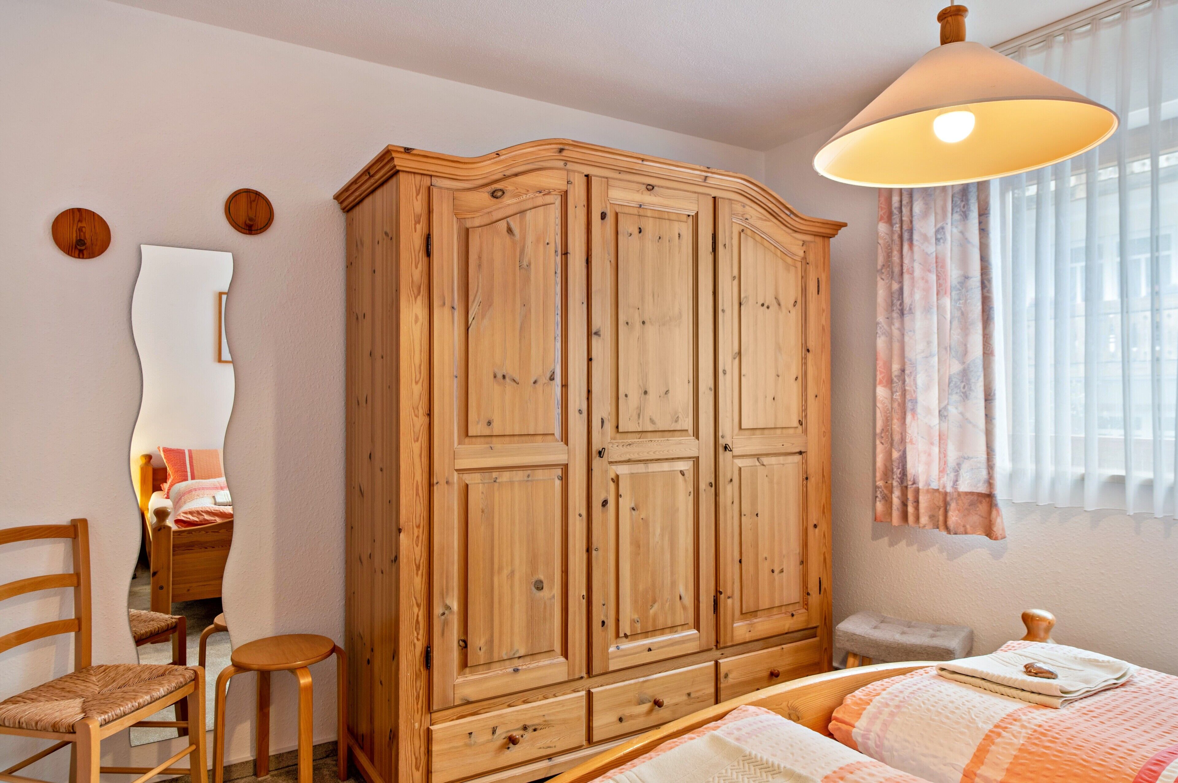 A bedroom with a wooden wardrobe, a bed, a chair, a small table, and a window with curtains.