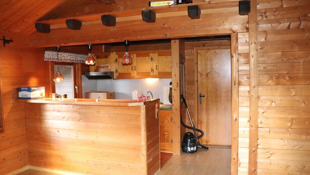 A wooden cabin interior with a kitchen area, wooden walls, and a door leading to another room.