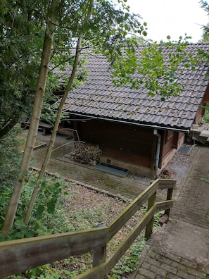 Ein traditionelles Holzgebäude mit Ziegeldach, einer Holztreppe und einem Gartenbereich mit Bäumen und einer Bank.