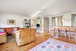 A living room with a sofa, wicker chair, wooden dining table, chairs, a bookshelf, and a patterned rug.