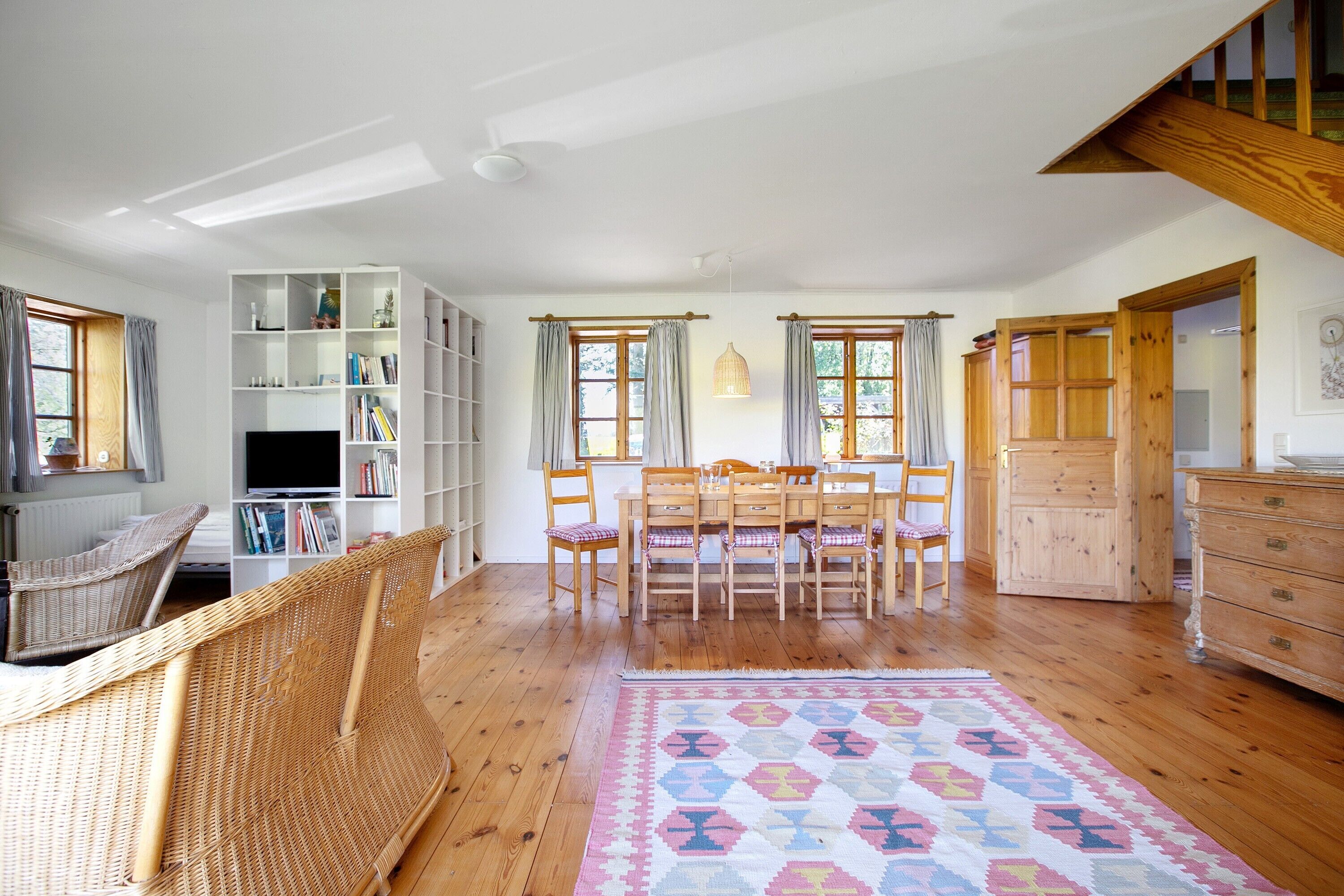 A spacious living room with a wooden floor, a dining table with chairs, a bookshelf, and a television.