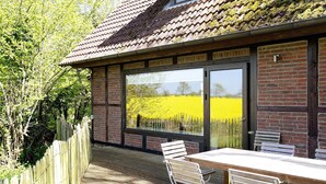 A patio with a table and chairs, a wooden fence, and a view of a yellow field through large windows. - Holiday home "Rosen" with terrace and garden in an idyllic location - relax and unwind! (Gelting)