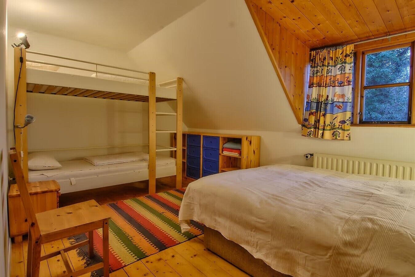 A room with a bunk bed, a single bed, a wooden desk, a radiator, and a window with curtains.