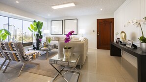 A modern living room with a sofa, armchair, coffee table, and large windows offering a city view. - Seaview balcony King Pacific Ocean Larcomar (Miraflores)