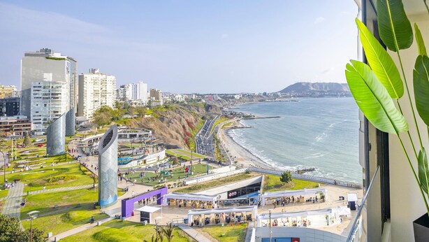 A coastal cityscape with modern buildings, a beach, and a view from a window with a plant. - Seaview balcony King Pacific Ocean Larcomar (Miraflores)