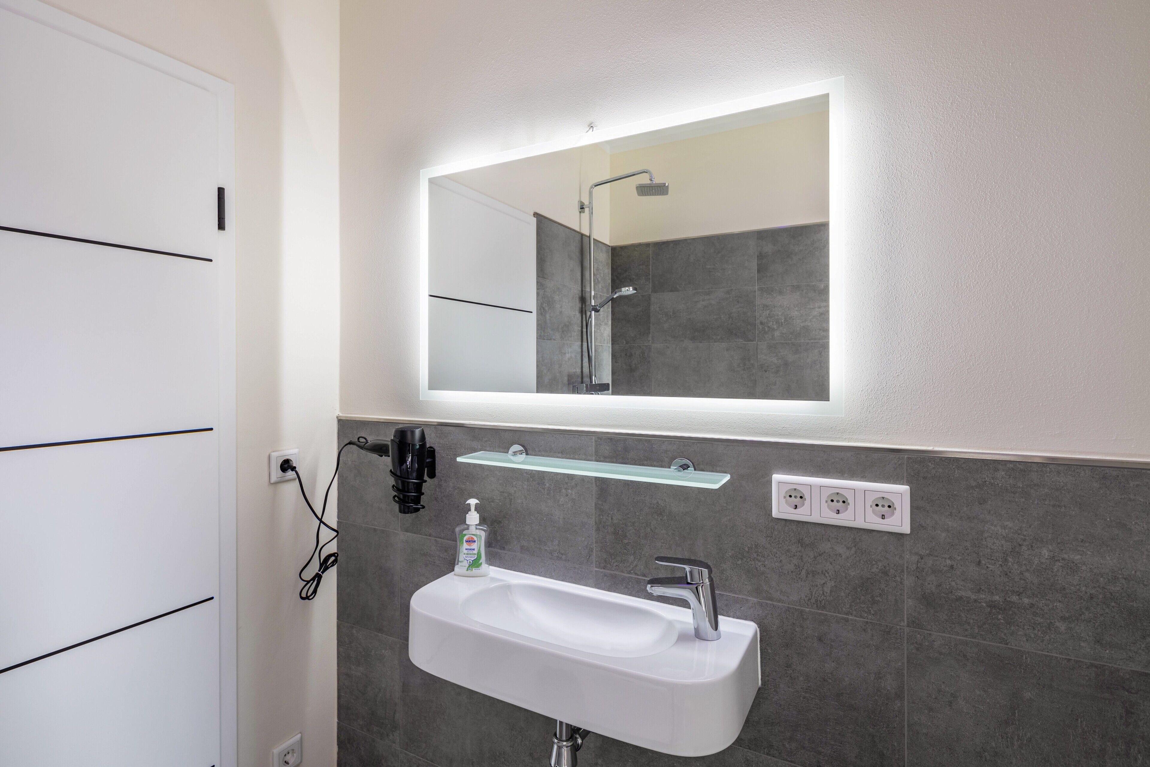 A bathroom with a white sink, a mirror, a shower, and a glass shelf.